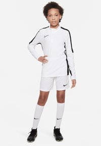 Camisa de manga larga blanca de Nike con detalles en negro, combinada con pantalones cortos blancos y calcetas blancas hasta la rodilla. Botas de fútbol negras completan el atuendo.