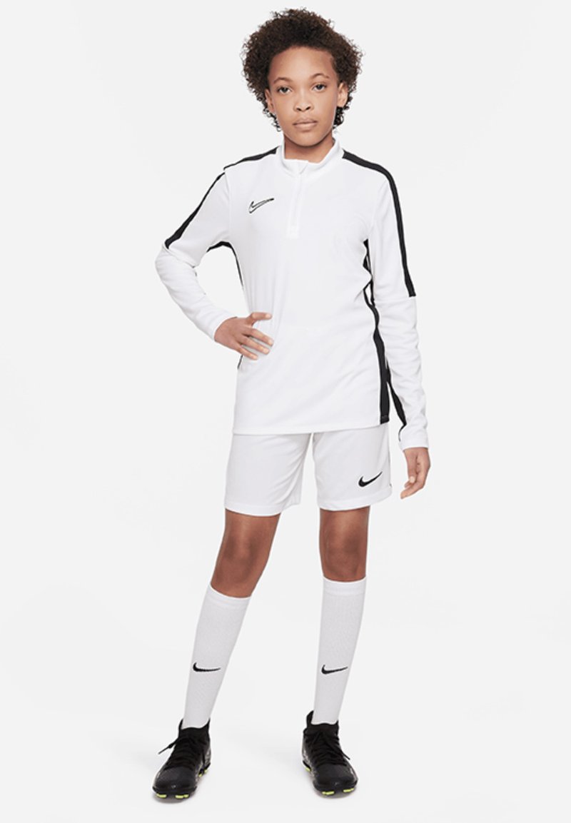 Camisa de manga larga blanca de Nike con detalles en negro, combinada con pantalones cortos blancos y calcetas blancas hasta la rodilla. Botas de fútbol negras completan el atuendo.