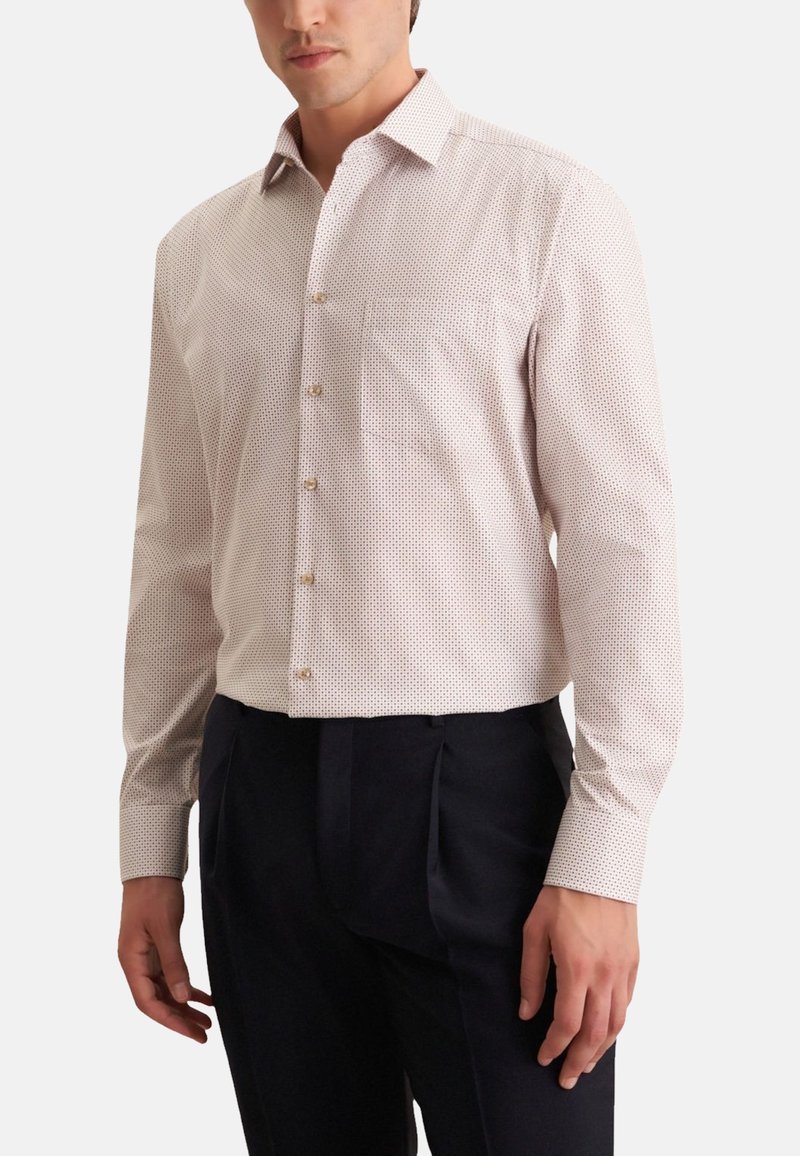 Camicia chiara a maniche lunghe con un piccolo motivo a pois, con colletto e taschino sul petto, abbinata a pantaloni scuri.