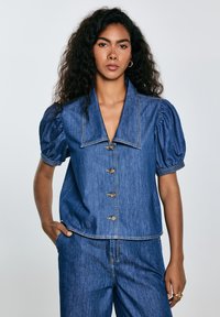 Denim-paita, jossa on puffihihat, suuri kaulus ja seitsemän nappia. Sininen väri, jossa näkyvät ompeluompelut ja rento istuvuus.