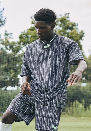 Giovane uomo in abbigliamento sportivo Puma con motivo nero che gioca a calcio all'aperto su erba, con una rete e alberi sullo sfondo.