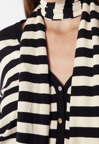 Gestreiftes Strickoberteil in Schwarz und Creme mit einem hohen Kragen. Verfügt über einen V-Ausschnitt und ein dekoratives Knopfdetail an der Brust, weiche Textur.