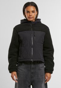 Schwarze Kapuzenjacke mit gestepptem Muster und samtigen Fleece-Ärmeln. Verfügt über einen Frontreißverschluss und Seitentaschen, kombiniert mit dunkelgrauen Hosen.