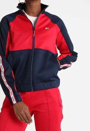 Persoon draagt een rood en marineblauw jack met ritssluiting en bijpassende rode broek, met merkstrepen langs de mouwen en broek.