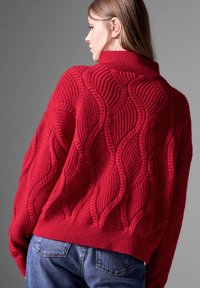 Roter Strickpullover mit einem strukturierten Wellenmuster, ausgestattet mit einem hohen Kragen und weiten Ärmeln. Das Material sieht dick und kuschelig aus.