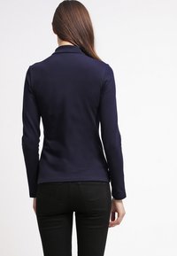 Blazer bleu marine à manches longues avec une coupe ajustée et des revers étroits. Tissu lisse, fente arrière unique et design minimaliste.