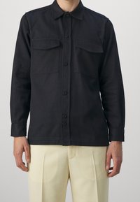 Black button-up shirt with two chest pockets, long sleeves, and a collar. Made of textured fabric, displayed with cream-colored pants.