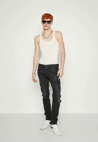 Λευκό αμάνικο μπλουζάκι, γκρι τρύπες τζιν και λευκά αθλητικά παπούτσια με μαύρες λεπτομέρειες. Το μοντέλο φοράει γυαλιά ηλίου και κολιέ, σε αντίθεση με έναν απλό φόντο.