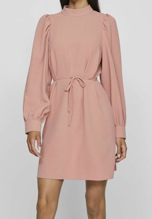 Frau trägt ein knielanges, staubrosa Kleid mit langen Puffärmeln, hohem Kragen und gebundenem Taillengürtel.