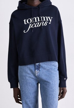 Osoba ubrana w granatową bluzę z kapturem Tommy Jeans oraz jasnoniebieskie dżinsy, stojąca na jednolitym tle.