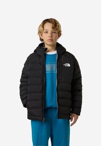 Μαύρο μπουφάν πάπλωμα με κουκούλα, με κλείσιμο με φερμουάρ και πλαϊνές τσέπες. Στο αριστερό στήθος υπάρχει λευκό λογότυπο. Φορεμένο πάνω από ένα ανοιχτό μπλε φούτερ.