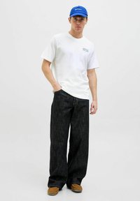 Camiseta blanca de manga corta con texto gráfico azul, combinada con jeans oscuros de corte amplio y calzado marrón. Diseño simple y casual.