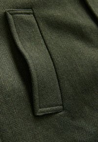 Dunkelgrüner Wollstoff mit einem Fischgrätmuster, das eine strukturierte Oberfläche und eine auffällige rechteckige Tasche aufweist.