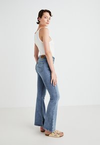 Lichtblauwe flared jeans van denim, met een hoge taille en een bruin leren label op de achterkant van de tailleband, gedragen met een wit tanktop.