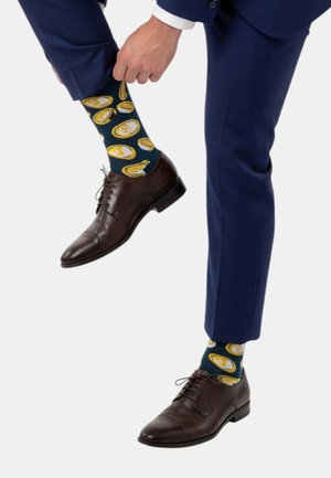 Mann in marineblauem Anzug, der bunte Socken mit gelben Münzmuster anpasst, trägt dunkelbraune Lederschuhe vor weißem Hintergrund.