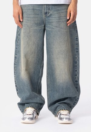 Person trägt locker sitzende, ausgewaschene blaue Jeans mit weiten Beinen und weiße Turnschuhe mit grauen und roten Details.