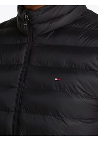 Czarna kurtka puchowa z poziomym pikowaniem, wysokim kołnierzem i przednim zamkiem błyskawicznym. Małe logo marki w pobliżu zamka w kolorach czerwonym, białym i niebieskim.