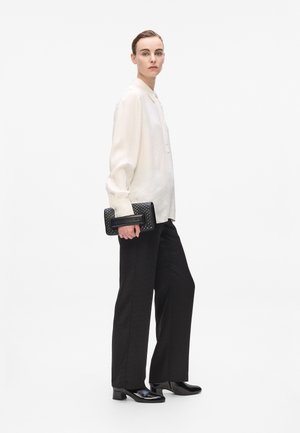 Black patterned trousers, white collared shirt, and a textured black clutch with detailing. Glossy black shoes complete the look.