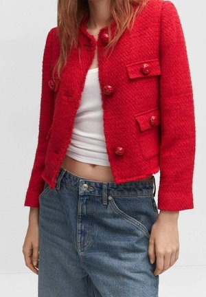 Femme portant une veste rouge texturée boutonnée sur un top court blanc et un jean bleu taille haute, debout devant un fond uni.