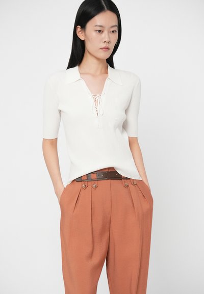 Ριγέ λευκό πόλο πουλόβερ με λαιμόκοψη με κορδόνια και κοντά μανίκια, συνδυασμένο με ψηλόμεσες, πλισέ παντελόνες σε χρώμα σκουριάς και καφέ ζώνη.