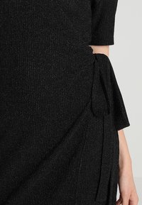 Schwarzes Kleid aus strukturiertem Rippstoff, mit einem seitlichen Knoten-Detail und kurzen Ärmeln. Das Material hat einen dezenten Schimmereffekt.