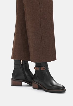 Stivaletti alla caviglia in pelle nera con cerniera laterale, cinturino marrone d'accento, tacco impilato e finitura liscia, abbinati a pantaloni marroni a quadri.