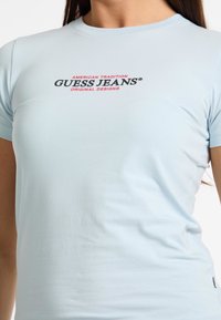 Camiseta de manga corta azul claro hecha de algodón, con un logo en rojo y negro que dice "AMERICAN TRADITION GUESS JEANS ORIGINAL DESIGNS."