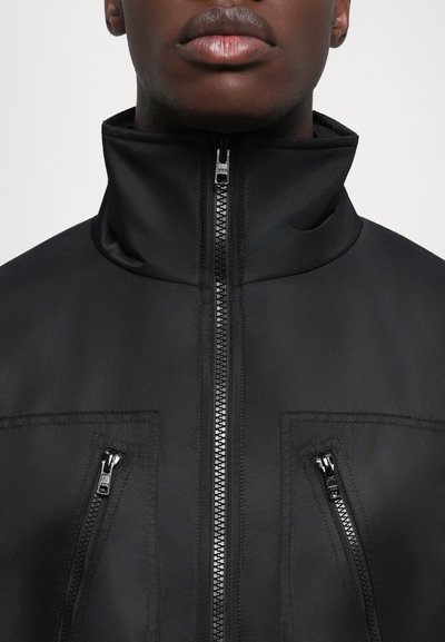 Svart jacka med hög krage och två zip-fickor framtill. Tyget verkar slätt med en subtil lyster och starka sömnadsdetaljer.