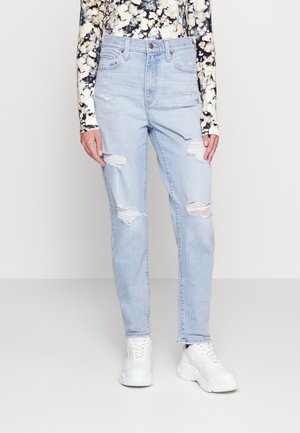 Personne portant un jean bleu clair déchiré, des baskets blanches épaisses et un haut à manches longues noir et blanc à motifs floraux, debout sur un fond uni.