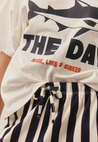 Valge T-särk musta shark graafikaga ja tekstiga "THE DAY"; koos siniste ja valgete triibuliste lühikeste pükstega, millel on paelael. 