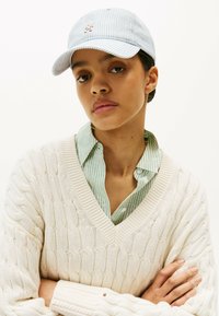 Cappellino a righe blu e bianche con logo frontale, abbinato a una camicia a righe verde e bianca sotto un maglione lavorato a maglia crema testurizzato.