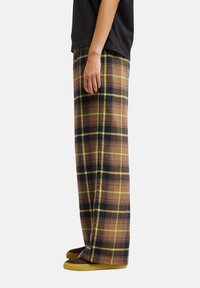 Personne portant un pantalon large à carreaux marron et noir, un haut noir et des chaussures marron à enfiler, debout de profil sur fond blanc.