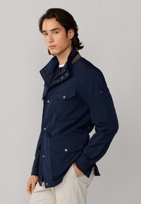 Chaqueta azul marino con cuello alto, dos bolsillos en el pecho y detalles contrastantes en los hombros. Textura suave y botones a presión visibles.