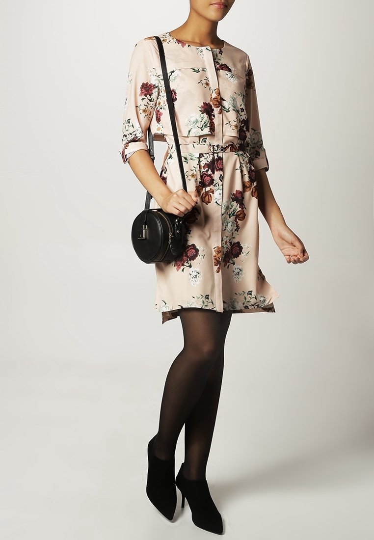 Femme portant une robe fleurie mi-longue avec ceinture, collants noirs, bottines à talons noires et un sac bandoulière rond noir.