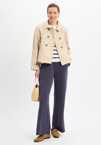 Beige dubbelgeknoopte blazer, witte trui met marineblauwe strepen, marineblauwe wijde broek, bruine loafer schoenen en een geweven beige tas. Lichte achtergrond.
