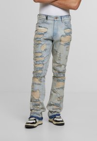 Šviesiai mėlynos džinsinės kelnės su keliais ištrintais lopais, suplėšytais kraštais ir tiesia kojų forma. Dėvimos su kontrastinėmis juodai baltomis sportbačiais.