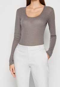 Kvinna klädd i en åtsittande grå långärmad topp med rund halsringning, instoppad i högmidjade vita skräddarsydda byxor, med en hand i fickan.