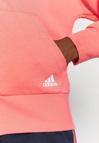 Felpa corallo con tasca frontale e polsini a coste. Presenta un logo Adidas bianco sul lato sinistro in basso. Texture morbida, design informale.
