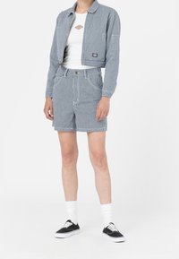 Blå och vit randig lättviktsjacka med dragkedja framtill, tillsammans med matchande shorts. Vit t-shirt under och svarta sneakers.