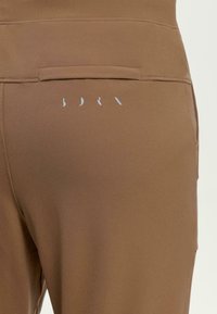 Brūni sporta bikses ar gludu tekstūru, kas aprīkotas ar mazu aizmugurējo kabatu un gaismas pelēku logotipu. Dizains ir pieguļošs ar augstu jostasvietu.