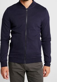 Mężczyzna w granatowej kurtce zapinanej na zamek z kołnierzem i bocznymi kieszeniami, w połączeniu z ciemnoszarymi spodniami, stojący na jednolitym tle.