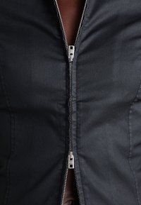 Sort zip-jakke med en glat, let blank overflade; har en sølvmetal lynlås fortil og et tætsiddende design.