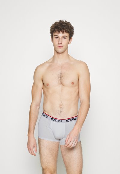 Young man standing against white background wearing light grey Moschino boxer briefs, facing forward with relaxed expression.