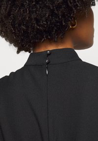 Svart tygplagg med hög krage och knäppning bak, med två tygklädda knappar och en slät yta.