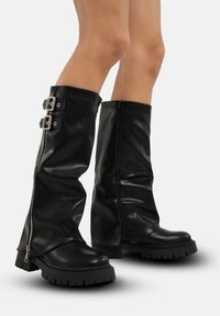 Botas negras hasta la rodilla hechas de cuero suave, que cuentan con dos hebillas decorativas y un cierre lateral, con suelas de plataforma gruesa y un acabado texturizado.