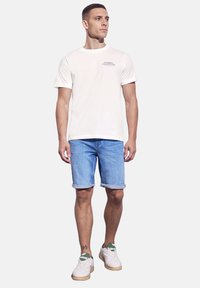 Bílý bavlněný tričko s krátkým rukávem a potiskem, v kombinaci s světle modrými džínovými šortkami. Model má na sobě bílé tenisky se zelenými detaily.