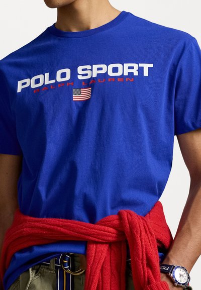 Camiseta de algodón azul real con la inscripción "POLO SPORT" en letras blancas y rojas y un gráfico de la bandera americana. Suéter rojo atado a la cintura.