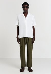 Chemise blanche à manches courtes et à boutons avec une finition texturée, associée à un pantalon cargo vert olive avec des poches latérales. Des sandales marron complètent le look.