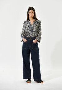 Blå skjorte med blomstermønster og knap-ned krave, parret med brede navy bukser med store lommer og kontrastsøm. Sandaler synlige.