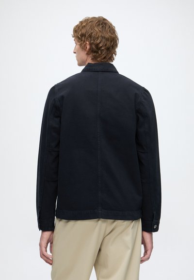 Homme aux cheveux bouclés portant une veste noire et un pantalon beige, debout dos à la caméra sur un fond blanc uni.